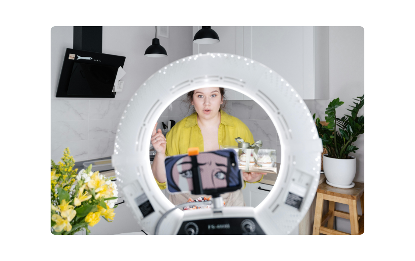 Woman using a ring light and phone to record a video in a kitchen, perfect for video creation side gigs.